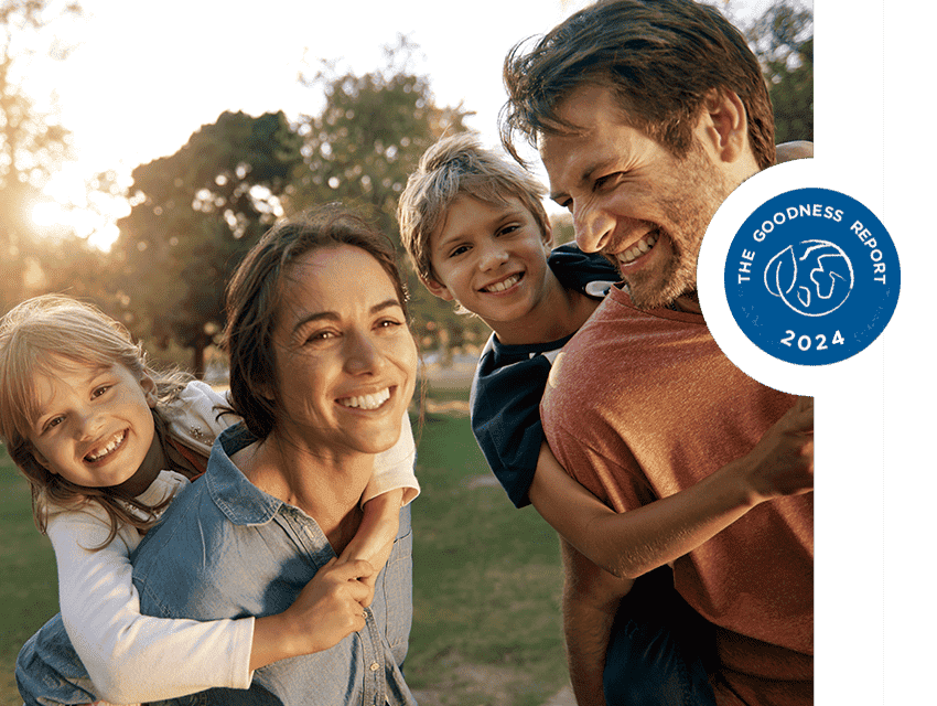 Happy family of four outdoors, with a mother and daughter and a father and son smiling and giving piggyback rides in a sunlit park setting. 
