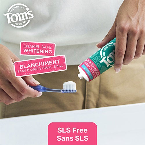 A person squeezing a pea-sized amount of white toothpaste from a Tom's of Maine tube onto a blue toothbrush. Graphic overlays emphasize "Enamel Safe Whitening" and "SLS Free" in English and French.